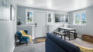 A modern living room with a blue sofa, a small table, and a chair. There are windows with views of trees and a door with a doormat.