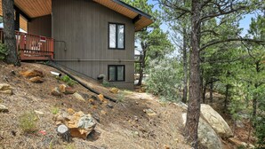 A modern two-story house with a wooden deck, situated on a sloped terrain with pine trees.