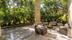 A covered outdoor lounge area with wicker furniture, a water feature, and lush greenery.