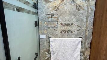 A bathroom with a glass shower enclosure, a marble-patterned wall, and a wooden door.