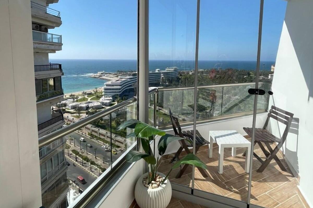 A balcony with a view of the ocean and cityscape, furnished with a chair, table, and potted plant.