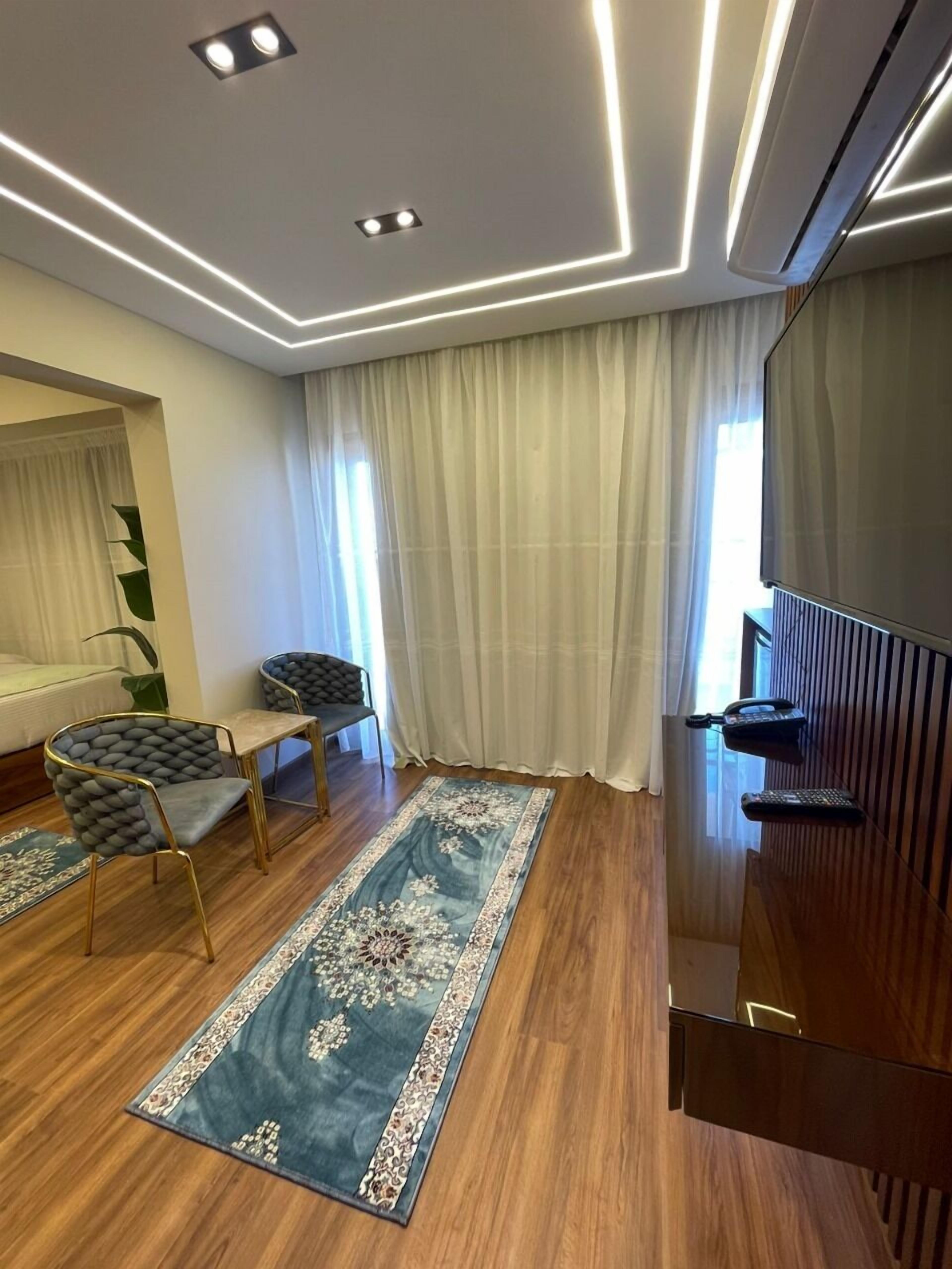 A modern hotel room with a wooden floor, a blue floral rug, a sofa, a chair, a small table, a mirror, and a large window with curtains.