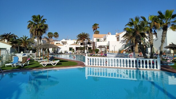 A swimming pool with lounge chairs and palm trees, surrounded by buildings with balconies. - Puerta del Sol, fully equipped apartment, ideally located close to all amenities (Caleta de Fuste)
