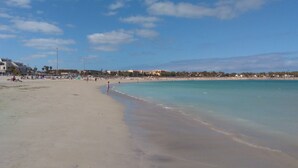 A wide sandy beach with clear turquoise water, a few people, and buildings in the background. - Puerta del Sol, fully equipped apartment, ideally located close to all amenities (Caleta de Fuste)