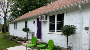 Une maison blanche avec un toit rouge, une porte violette et un petit jardin avec des chaises vertes et des plantes.
