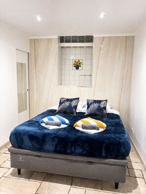 Una cama con cubierta azul oscuro, dos almohadas con motivos de hojas y un florero con girasoles en la pared.