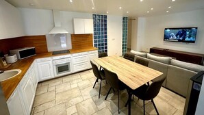 A modern kitchen with white cabinets, a wooden countertop, and a built-in oven. There is a dining area with a wooden table and chairs, and a TV mounted on the wall.