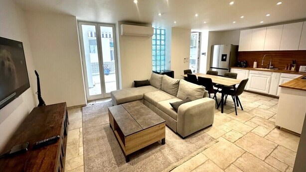 A modern living room with a sectional sofa, a coffee table, and a TV. There is a kitchen area with white cabinets and a dining table.