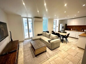 A modern living room with a sectional sofa, a coffee table, and a TV. There is a kitchen area with white cabinets and a dining table.
