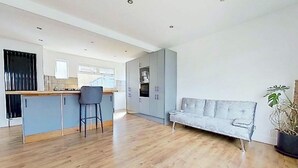 A spacious living area with a wooden floor, a grey sofa, a bar area with stools, and a kitchenette in the background. - South Downs Retreat (Sompting)