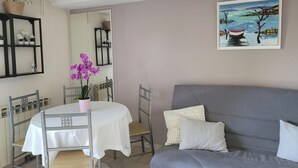 A dining area with a table and chairs, a gray sofa with pillows, a painting on the wall, and shelves with plants and decorative items. - Charming 35 m2 apartment with terrace and garden (Domont)