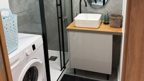 A compact bathroom with a glass-enclosed shower, a sink with a mirror, a washing machine, and a small storage area.