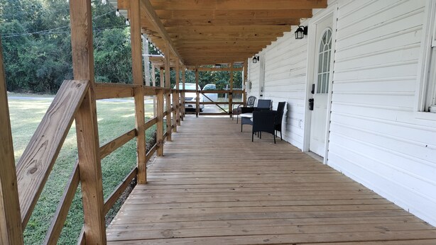 Une terrasse en bois avec une rambarde, un espace salon couvert avec des chaises, et un bâtiment blanc en arrière-plan.
