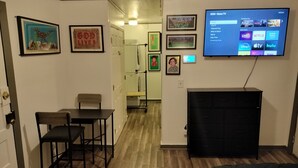 Un espace de vie compact comprenant un coin cuisine, un espace repas avec une table et des chaises, un lit et un téléviseur fixé au mur, qui affiche l’interface Roku.
