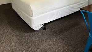 A hotel room with a bed, a ceiling fan, a wall-mounted coat rack, a desk with a chair, and a decorative wall piece.