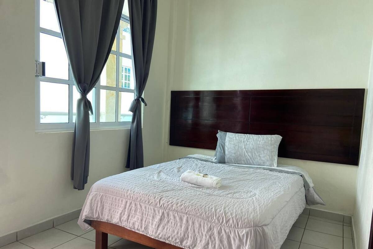 A neatly made bed with a white comforter and a folded towel, a wooden headboard, and a window with curtains.