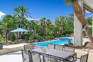 Zona junto a la piscina con mesa y sillas, rodeada de palmeras y un cielo azul despejado.