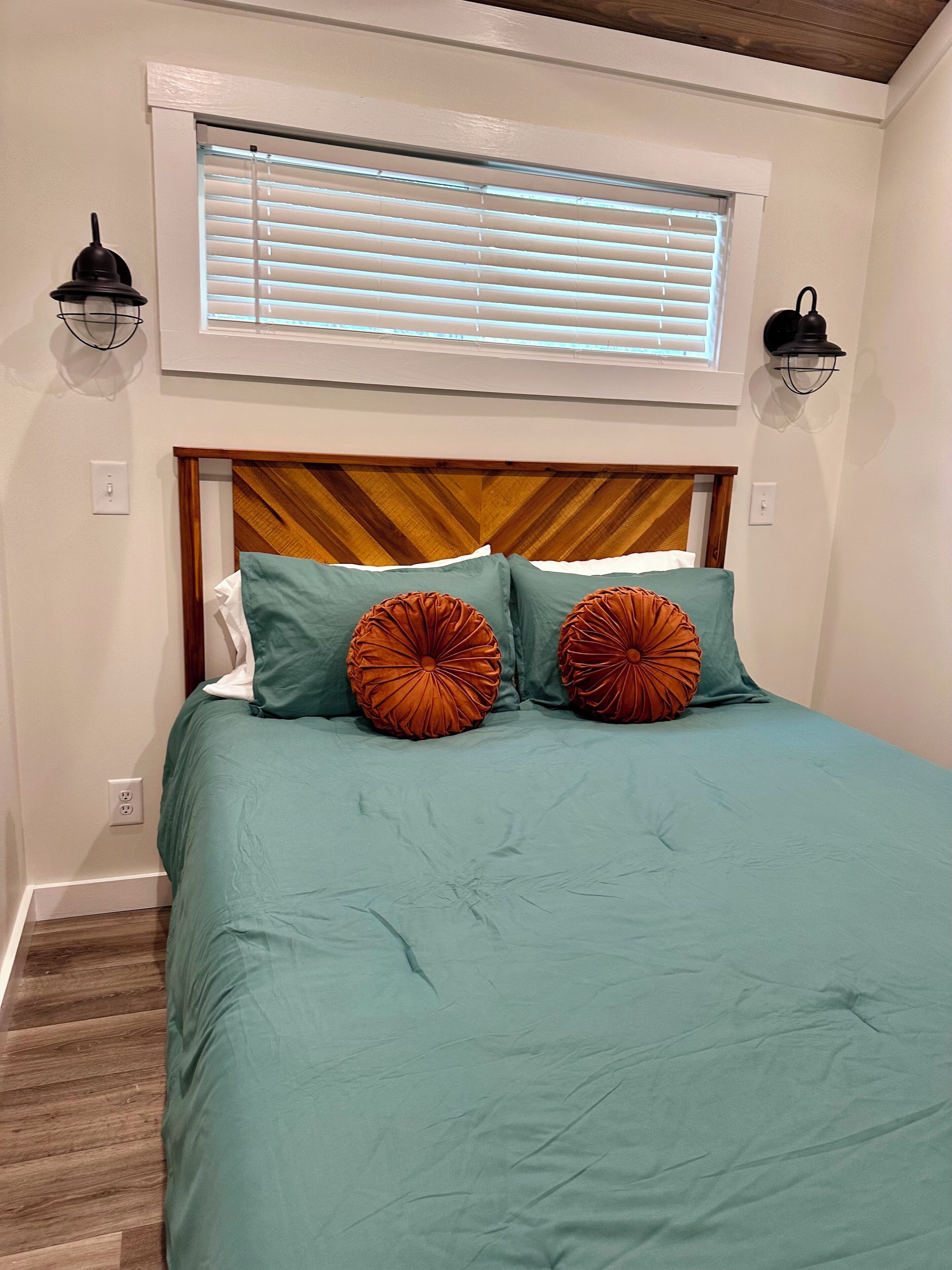 Un dormitorio con cabecera de madera, ropa de cama color verde azulado y dos almohadas decorativas anaranjadas.