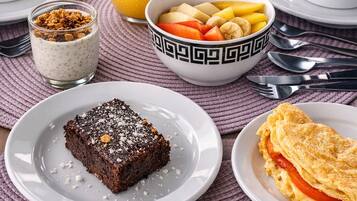 Un desayuno con un vaso de zumo de naranja, un bol de fruta, una porción de pastel, un vaso de leche y un plato de huevos revueltos.