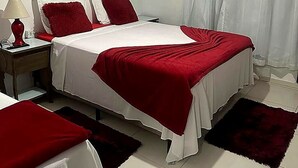 A bedroom with a bed, a nightstand, a lamp, a window with curtains, and an air conditioning unit. - Pousada Rainha do Mar  (São Francisco do Sul)