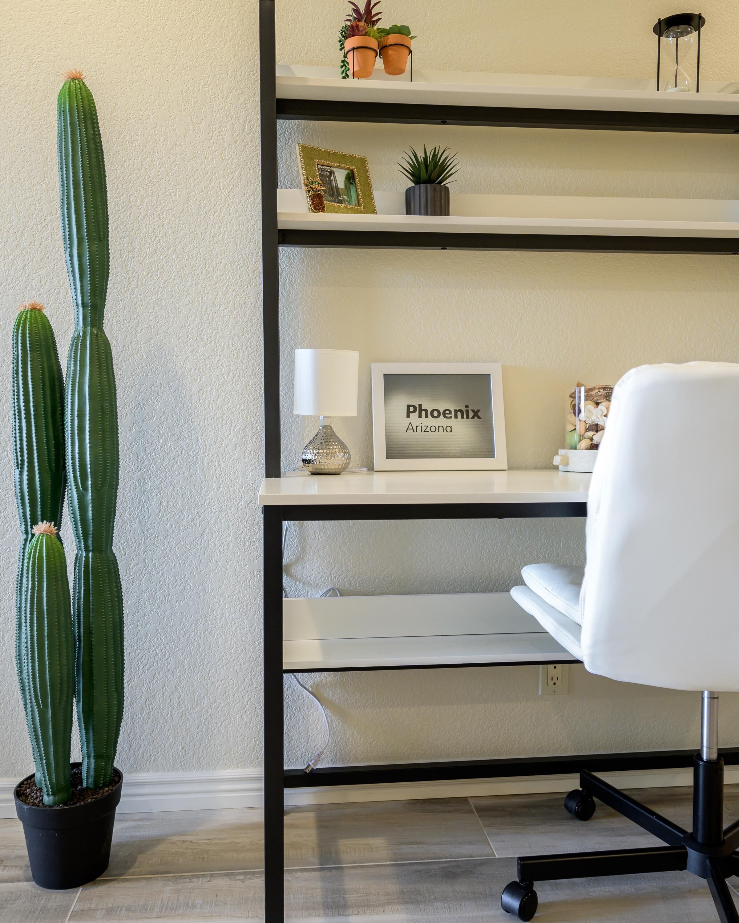 A workspace with a cactus, a framed sign reading 'Phoenix Arizona,' a lamp, and a white office chair.