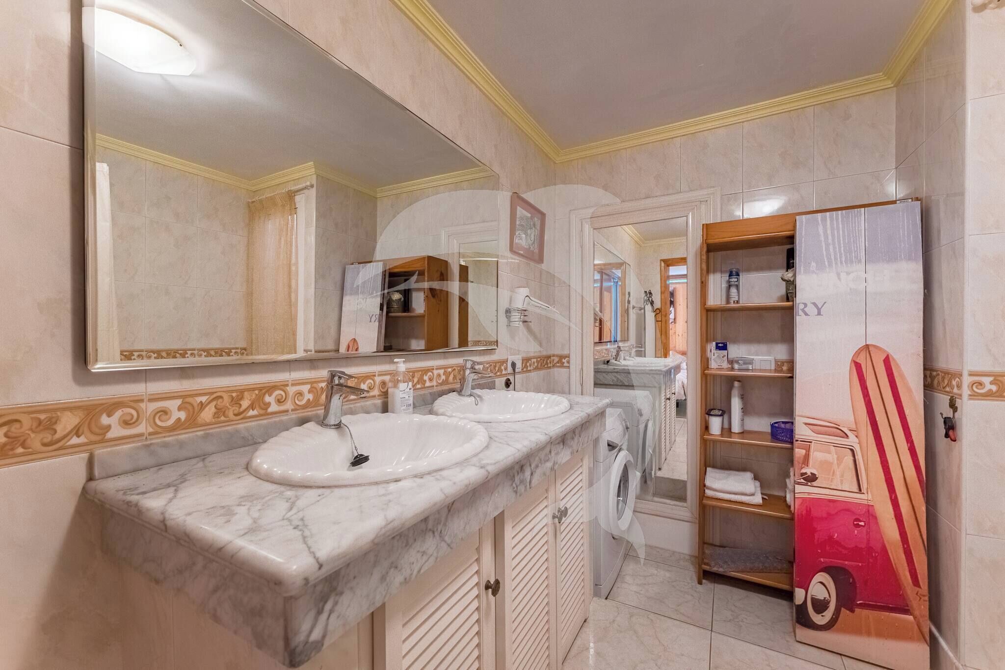 A bathroom with two sinks, a large mirror, a washing machine, and a shelf with toiletries.