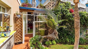 A patio area with a glass door leading to a room, surrounded by lush greenery and a palm tree. - Relax in El Médano's Hidden Gem 1-bedroom Apartment (Granadilla de Abona)