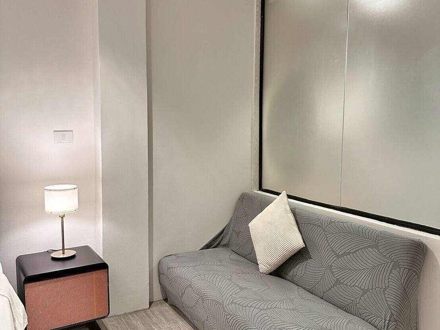 Ein modernes, minimalistisches Zimmer mit einem grauen Sofa, einem kleinen Beistelltisch mit Lampe und einem großen Fenster.