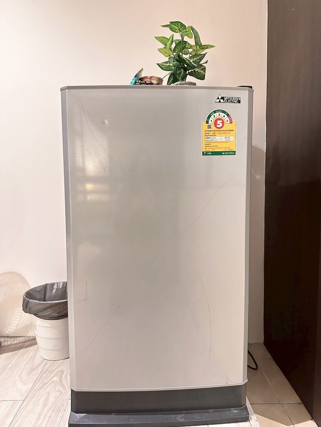 Un réfrigérateur blanc orné d’un autocollant Mitsubishi Electric, avec une petite plante posée sur le dessus et une poubelle à gauche.