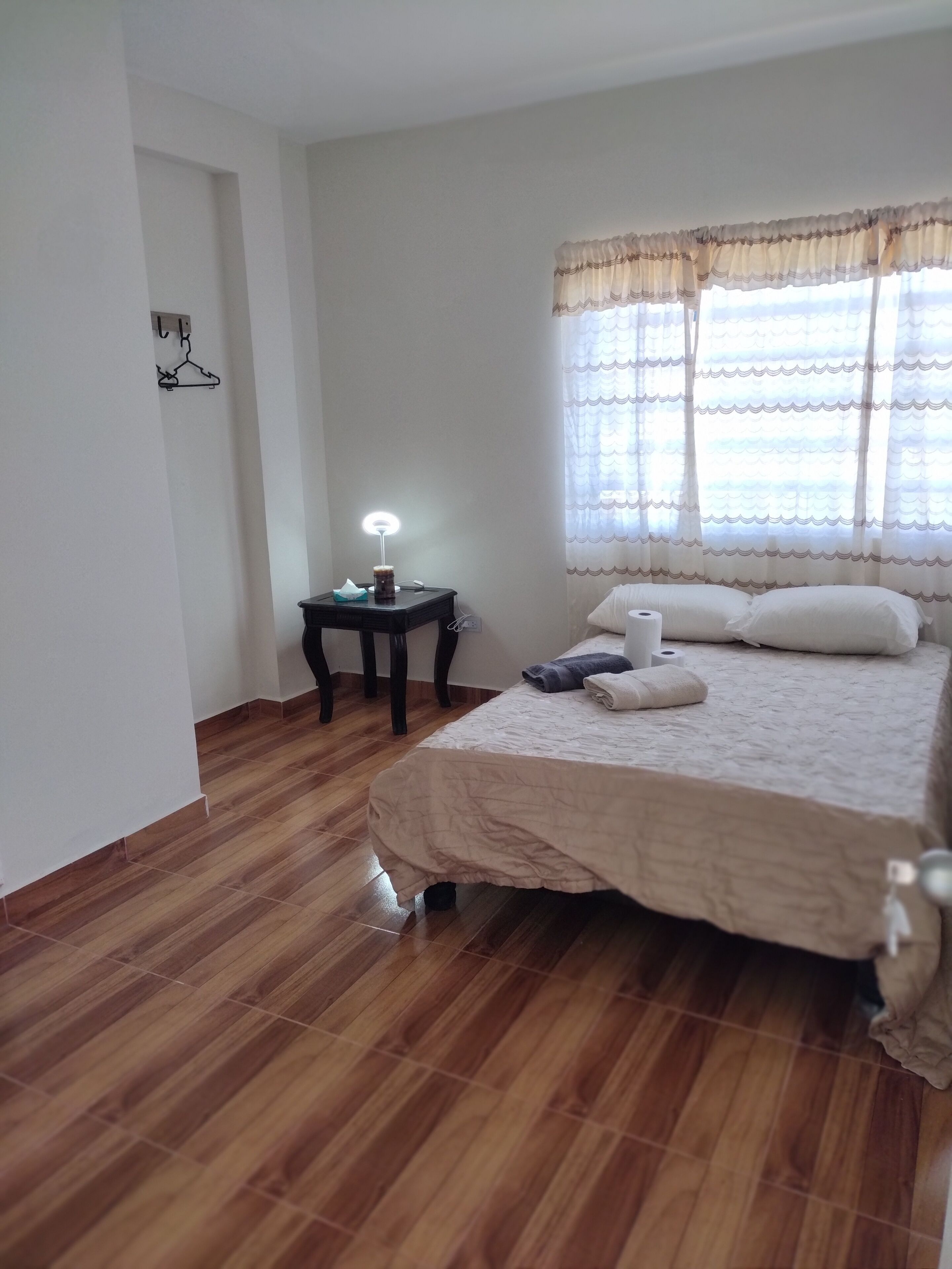 A bedroom with a wooden floor, a bed with pillows and a blanket, a bedside table with a lamp, and a window with sheer curtains.