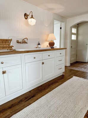 A white bathroom with a wooden vanity, a wall-mounted light fixture, and a white rug on the floor. - 1920s cottage with AC in charming Greenleaf (Greenleaf)