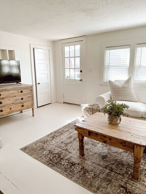 A room with a wooden coffee table, a white sofa, a patterned rug, a wooden dresser, and a television. - 1920s cottage with AC in charming Greenleaf (Greenleaf)