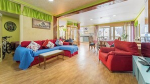 Ein großzügiges Wohnzimmer mit einem roten Sofa, einem blauen Sessel und einem Holztisch. Im Hintergrund ist ein Essbereich mit einem roten Stuhl und Tisch zu sehen.