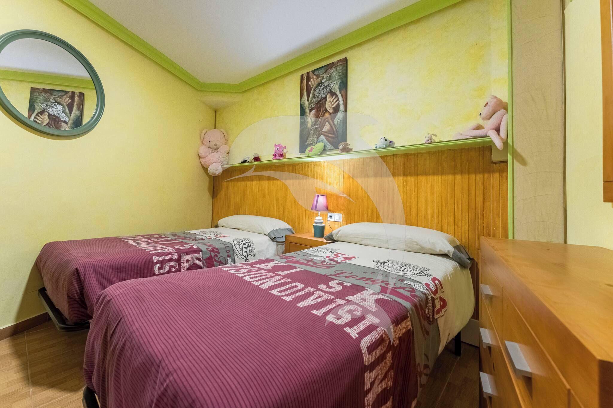 A room with two beds, a wooden dresser, a mirror, and a shelf with decorative items.