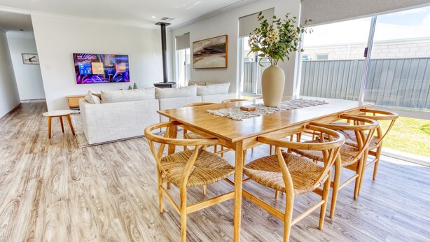 A modern living room with a wooden dining table, wicker chairs, a white sofa, and a large window with a view of the backyard.