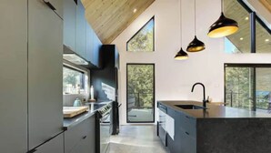 A modern kitchen with dark cabinetry, a central island, pendant lighting, and large windows offering views of the outdoors. - Chalet OSLO | Design & forest | Spa - Silence (Saint-Cã´Me)