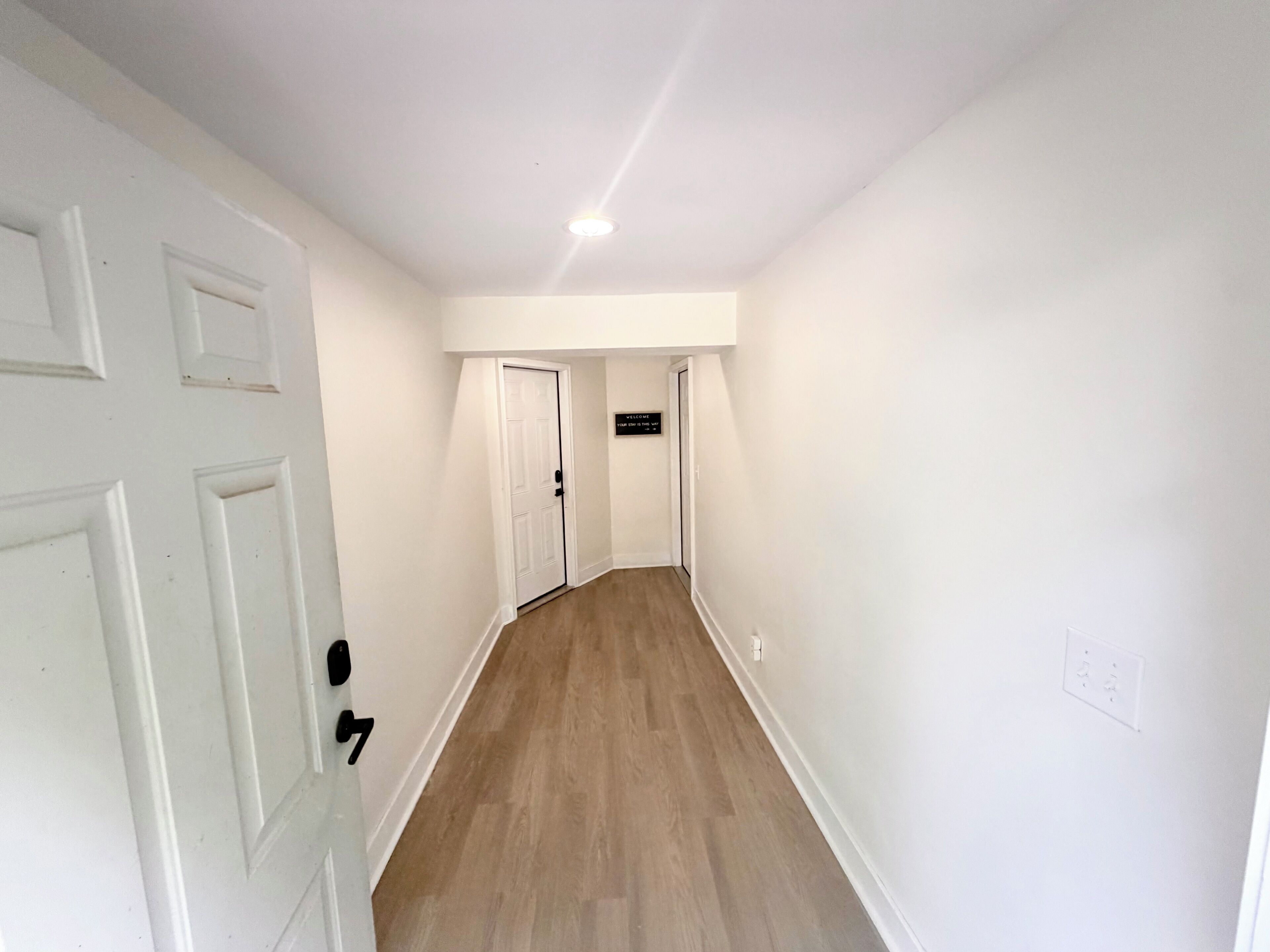A hallway with white walls and wooden flooring, leading to a door with a sign above it.