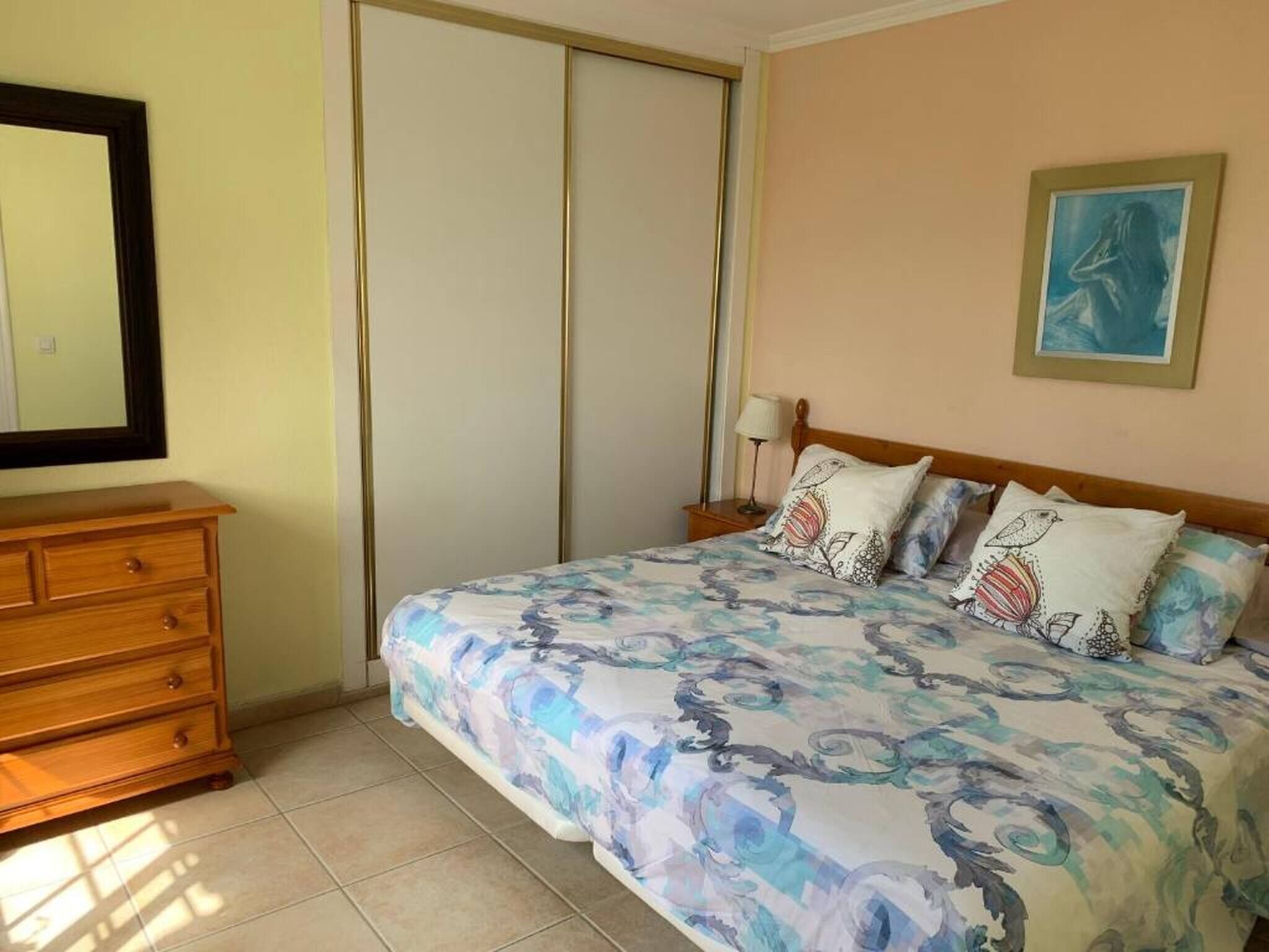 A bedroom with a bed, a wooden dresser, a nightstand, a lamp, and a painting on the wall.