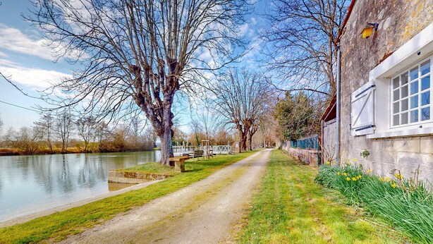 Un sentier étroit bordant une rivière, avec des arbres sans feuilles de chaque côté et un bâtiment doté d’une fenêtre.