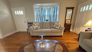 Ein Wohnzimmer mit Holzboden, einem beigefarbenen Sofa, einem Glastisch und einem Eckfenster mit Vorhängen.