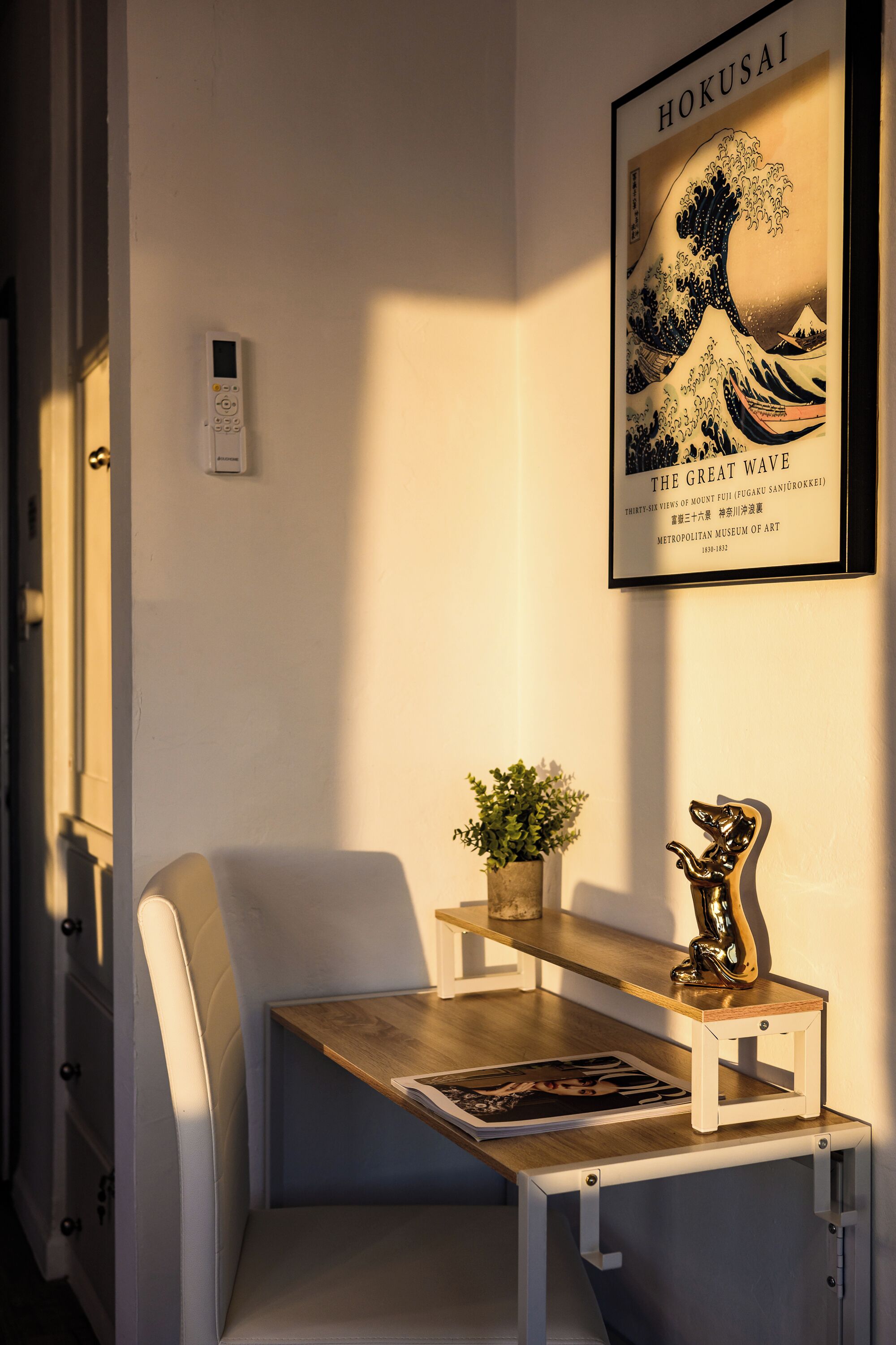 Une pièce avec une chaise blanche, une petite table sur laquelle se trouve une plante et un objet décoratif, une œuvre d’art encadrée accrochée au mur et un thermostat.