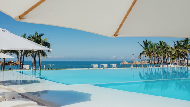 Une piscine d’un bleu éclatant, bordée de chaises longues et de parasols blancs, avec des palmiers et une vue sur l’océan.