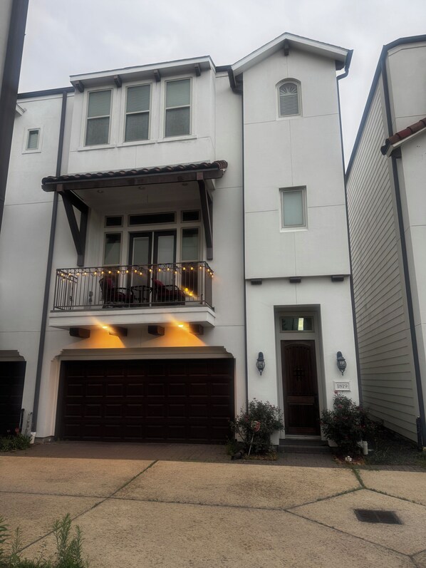 A two-story residential building with a garage, a balcony with string lights, and a small garden area.