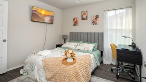 Un dormitorio con una cama, un escritorio y una silla. Hay un cuadro en la pared y una ventana con cortinas.