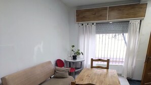 A living room with a wooden sofa, a dining table with chairs, a small round table with a potted plant, and a window with curtains. - The House of Napoli (Trelew)