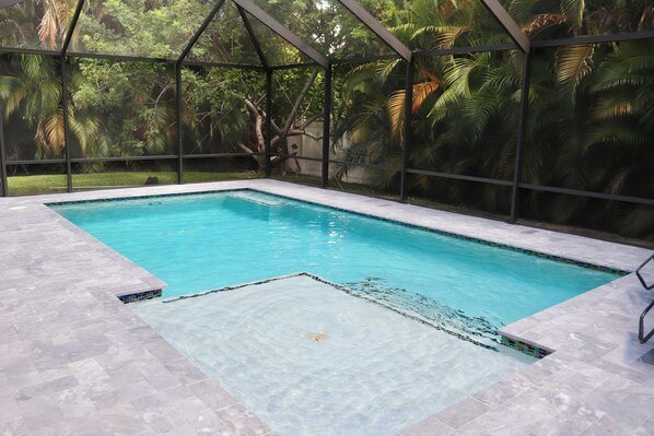 Una piscina interior rectangular con agua azul cristalina, rodeada por una zona cubierta con estructura metálica y paredes de vidrio.
