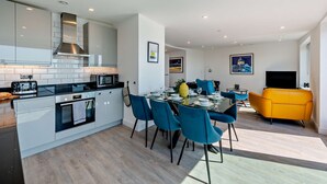 A modern kitchen with a dining area, featuring a central island, blue chairs, and a yellow sofa. - Penthouse on the Seafront (Worthing)
