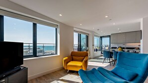 A modern living room with a large TV, a yellow armchair, and a blue armchair. There is a dining area with a table and chairs, and a kitchen with cabinets and a window. - Penthouse on the Seafront (Worthing)