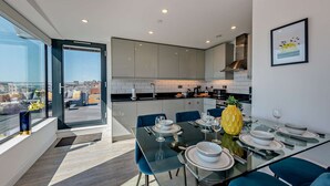 A modern kitchen with a dining area, featuring a glass table set for four, blue chairs, and a city view through large windows. - Penthouse on the Seafront (Worthing)