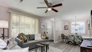 A modern living room with a gray sofa, a dining table with chairs, a ceiling fan, and large windows with blinds. - Modern living for visit DC/UMD, 2 bedrooms, 4 beds with central AC/heating (Hyattsville)
